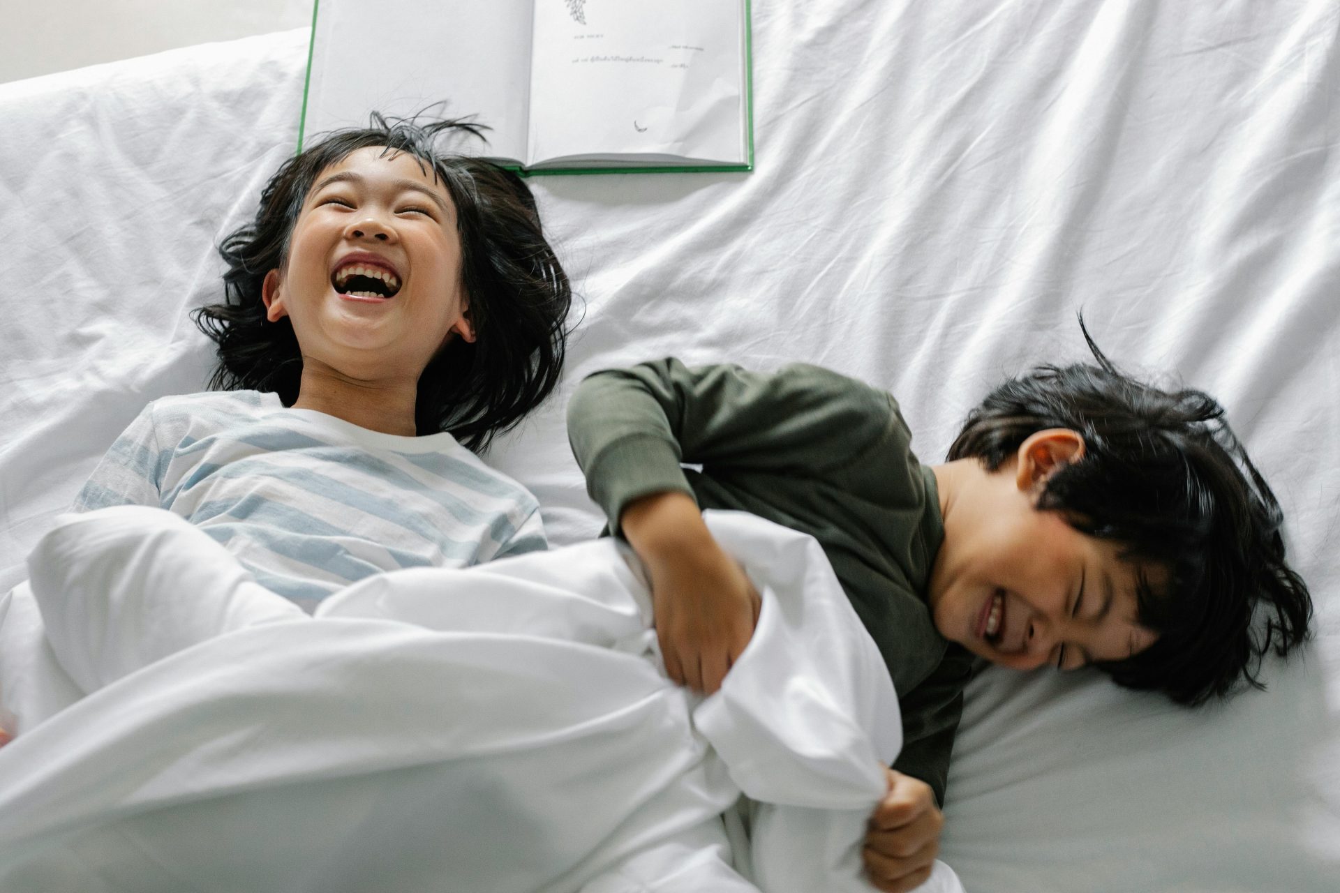 Two happy children laughing together on a cozy bed in the morning light.