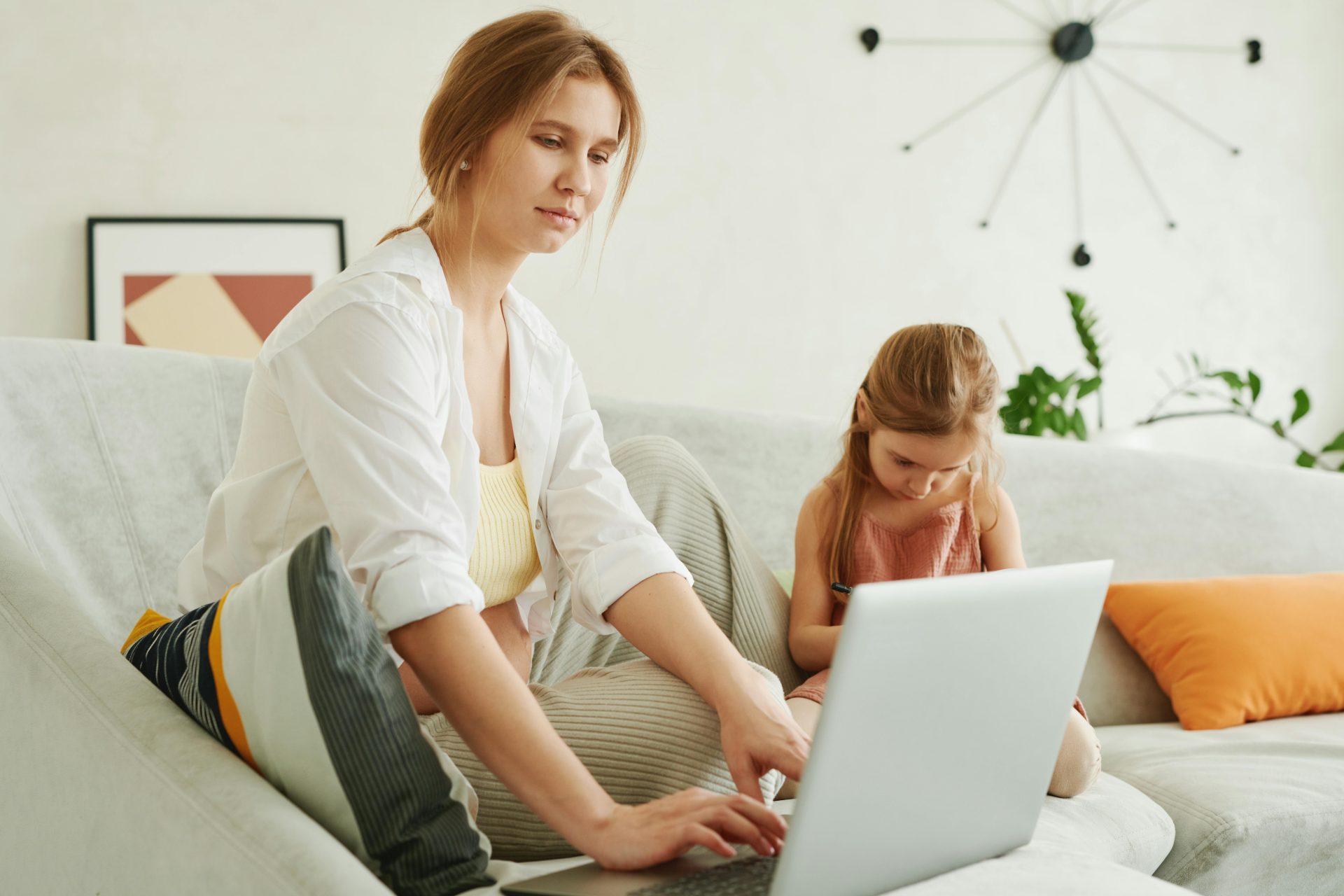 A mother and daughter use a laptop together on a cozy sofa indoors, multitasking family life.